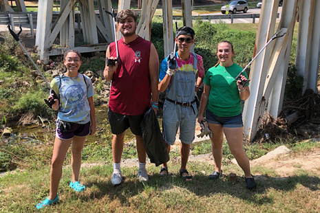 Four students stand in front of a metal beam bridge with trash collector poles.