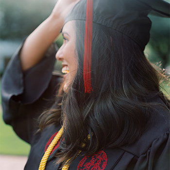 Student wearing graduation cap and gown adjusting her hat smiling in the sun.