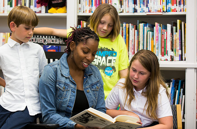 Student reading to kids with a shelf of books in the background.