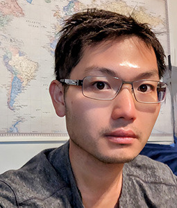 man sitting in front a of a world map with short brown hair and square rimmed glasses on