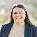 a woman with long dark hair and a dark blue jacket smiling outside