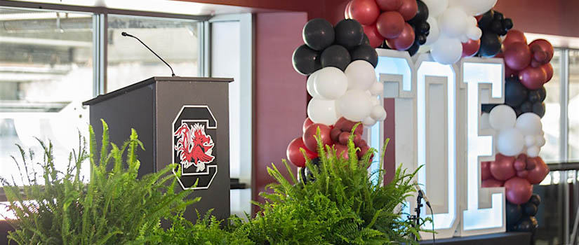 A festive podium decorated with the USC logo in the left side, and an aerch of black and garner balloons at the right.