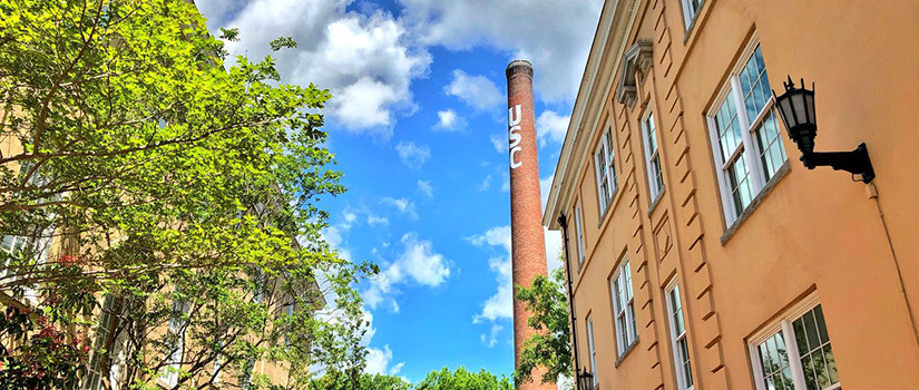 The UofSC water tower next to campus buildings and tress with a view of blue sky above with clouds. 