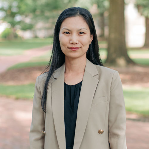 Smiling woman with long black hair, wearing a black dress shirt and beige suit jacket