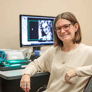 Student Kate Kuisel wearing glasses and tan top sits in front of a microfilm machine and computer screen