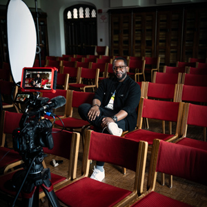 Camera films Jabari Evans while he sits in a room full of red chairs. 