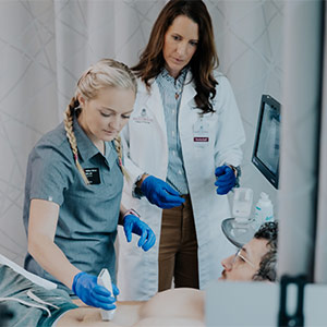 two women nurses stand at hospital bedside, one using a portable ultrasound on a patient