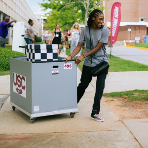 student in gray shirt pulling gray box of belongings on sidewalk during move-in