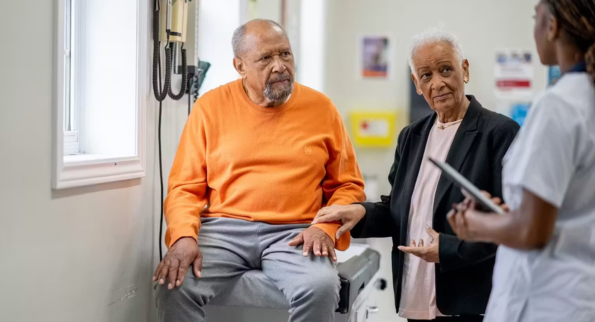 Older man sits on an exam table, older woman by his side, their doctor stands facing them.